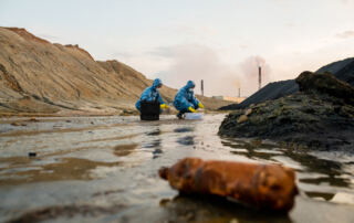 Two ecologists in rubber gloves, protective coveralls, eyeglasses and respirators studying characteristics of toxic water in dangerous area